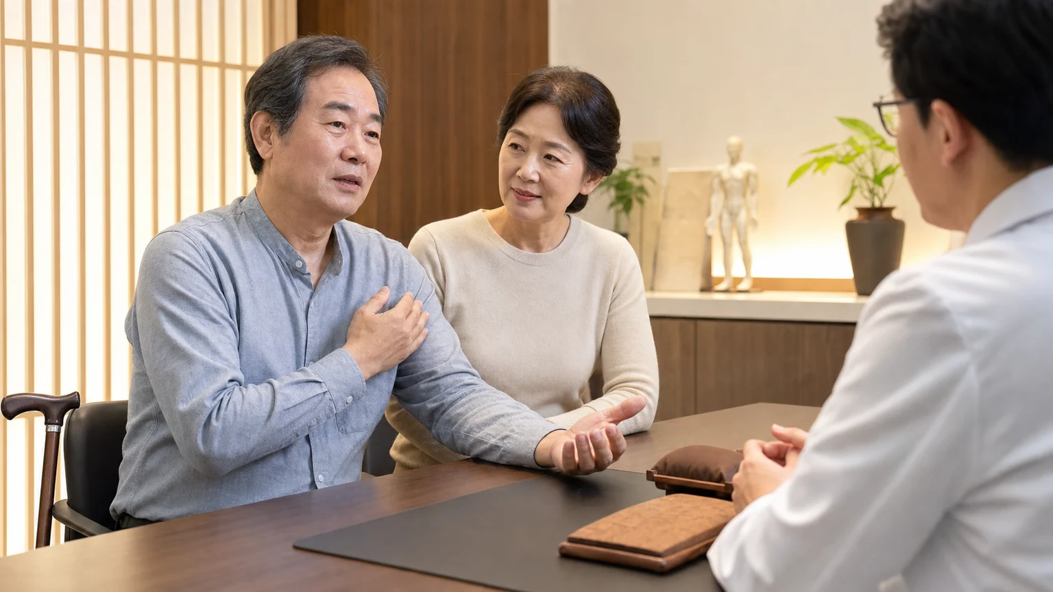 한국인 뇌졸중 후유증 환자와 보호자가 국내 한의원 상담실에서 남은 편마비와 생활 불편을 설명하는 장면